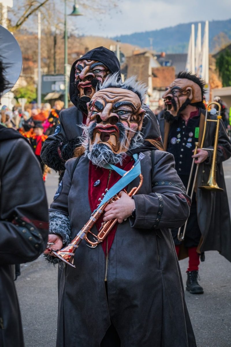 carnival, musician, trumpet, trombone, wooden mask, black coat, beard, fake mustache, costume, parade, region, traditional, public event, band, festival, colorful, face paint, outdoor event, vibrant festival