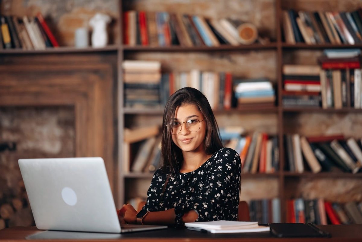 library, computer, glasses, desk, books on shelf, education, study, technology, laptop, student, sitting, smiling, female, office, knowledge, concentration, decoration, home library, professionalism, furniture, online