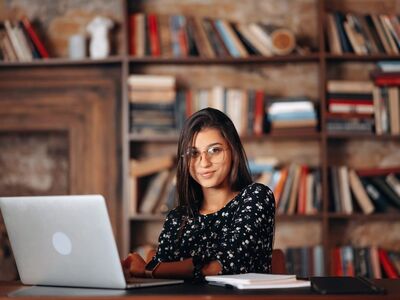 library, computer, glasses, desk, books on shelf, education, study, technology, laptop, student, sitting, smiling, female, office, knowledge, concentration, decoration, home library, professionalism, furniture, online